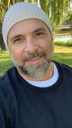 a man with a beard wearing a beanie smiles at the camera while sitting in a park