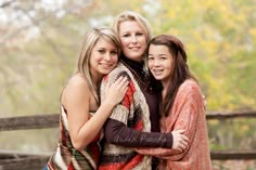 three women hugging each other while standing in front of a wooden fence with trees behind them