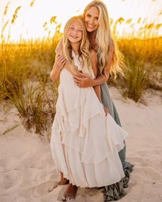 two women hugging on the beach in front of some tall grass and sand with sea oats behind them