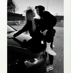 a man and woman sitting on the hood of a car kissing each other in black and white