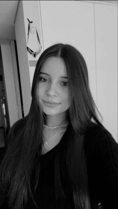 a young woman with long hair wearing a black shirt and silver necklace standing in front of a white wall