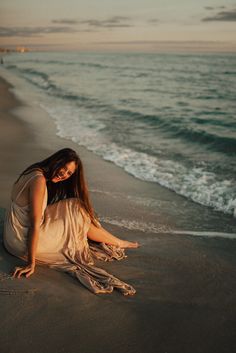 a woman sitting on top of a sandy beach next to the ocean with her hair blowing in the wind