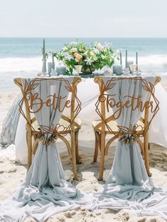 two chairs sitting on top of a sandy beach next to a table covered in white cloths