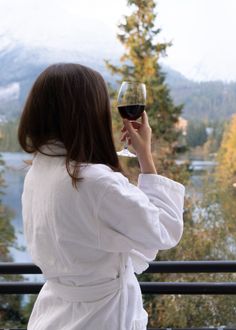 a woman holding a glass of wine looking out over a lake and mountains in the background