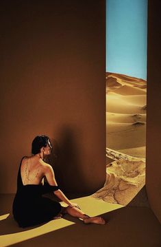 a woman sitting on the ground in front of a desert scene with sand dunes and blue sky