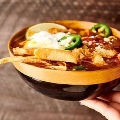 a person holding a bowl of chili and tortilla soup with sour cream on top