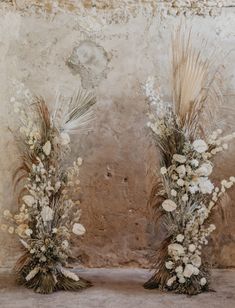 two vases with dried flowers and grasses in them against a stone wall, side by side