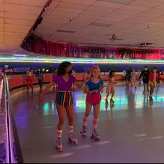 two girls on roller skates in an indoor skating rink with lights and people walking around