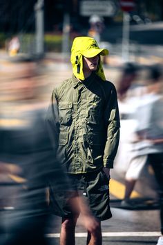 a man wearing a yellow hat and green jacket walking down the street with his hands in his pockets