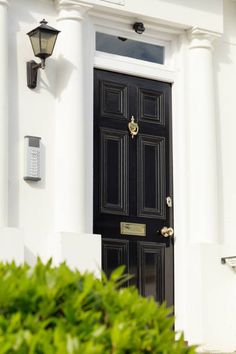 a black door is in front of a white building with green bushes and two lights