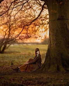 a woman sitting on the ground next to a tree talking on a cell phone while wearing a long brown dress