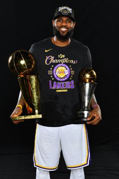 a man holding two trophies and wearing a black t - shirt with the lakers logo on it