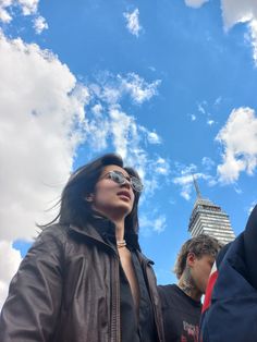 a woman wearing sunglasses standing in front of a tall building under a cloudy blue sky