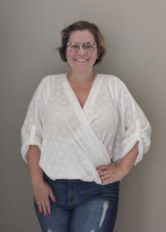 a woman with glasses standing in front of a wall wearing jeans and a white blouse