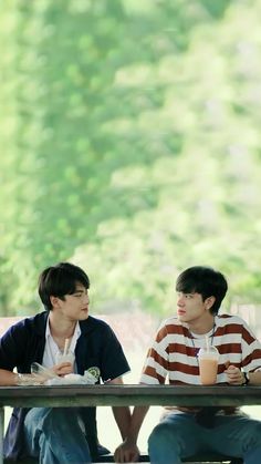 two young men sitting at a picnic table talking to each other, with trees in the background