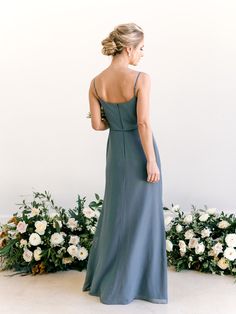 a woman standing in front of flowers wearing a long blue dress with spaghetti straps and low back