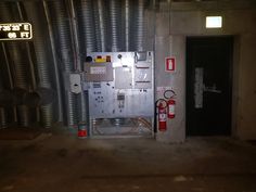 an electrical panel in the middle of a room with metal pipes on either side and red fire extinguishers at the end