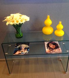 a glass table topped with two vases filled with flowers next to magazines on top of a wooden floor