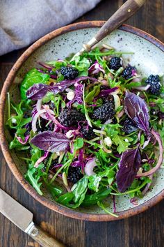 a bowl filled with greens and blackberries on top of a wooden table next to a knife