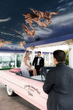 a bride and groom standing in front of a pink car with cherubs painted on the ceiling