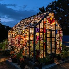 a glass house is lit up at night with flowers on the side and potted plants in front