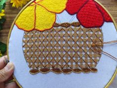 a person holding up a small embroidery project with yellow and red flowers in the background