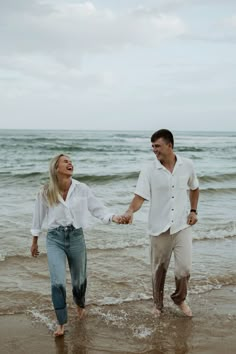 a man and woman are holding hands while walking in the water at the beach on a cloudy day