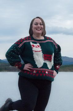 a woman standing in front of a body of water wearing a sweater with an image of santa clause on it