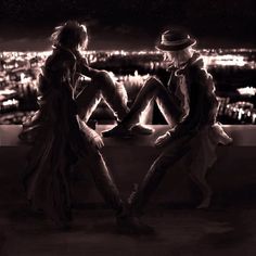 two people sitting on a ledge in front of a cityscape at night, one man is wearing a hat and the other has his legs crossed