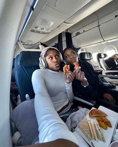 two women sitting in the back of an airplane eating sandwiches and pretzels with headphones on