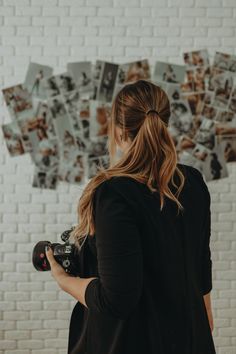 a woman holding a camera in front of a white brick wall with pictures on it