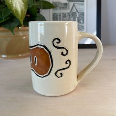 a white coffee mug with a brown and black design on the side sitting on a table next to a potted plant