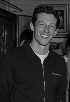 black and white photograph of smiling man in room with framed pictures on wall behind him