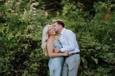 a man and woman standing next to each other in front of some green trees with their arms around each other