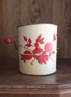 an old tin can with red flowers painted on it sitting on a wooden table next to a wall