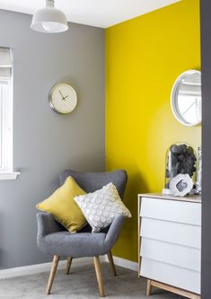 a living room with yellow and gray walls, grey chair, white dresser and clock on the wall