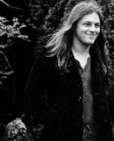 a black and white photo of a young man in front of some trees with long hair