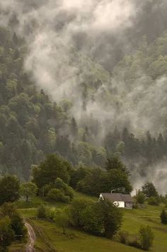 a house in the middle of a lush green hillside surrounded by fog and low lying clouds