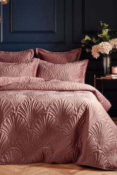 a bed with pink comforter and pillows in a room next to a vase filled with flowers