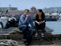 a man and woman sitting on top of a dock next to the ocean with boats in the background