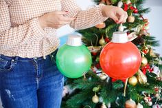 a woman holding two christmas ornaments in front of a christmas tree with decorations on it