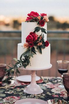 a white cake with red flowers on top sitting on a table next to wine glasses