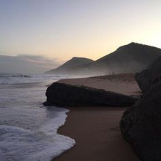 the sun is setting at the beach with waves crashing on the shore and mountains in the distance