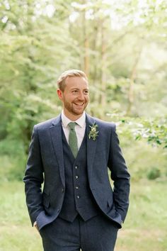 a man wearing a suit and tie standing in front of some trees with his hands in his pockets