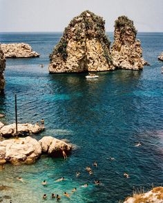 people are swimming in the water near some large rocks and small boats on the ocean