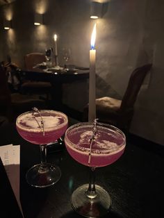 two glasses with pink liquid sitting on a table next to a lit candle in a dark room