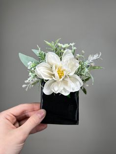 a hand holding a small black box with white flowers in it and greenery on top