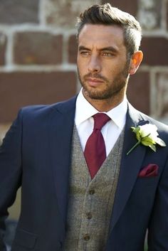 a man wearing a suit and tie with a flower in his lapel, standing next to a brick wall