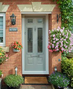 a grey front door surrounded by potted plants and flowers on the side of a brick building