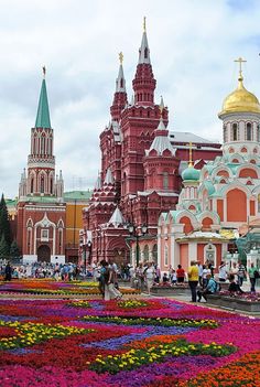 many people are walking around in front of some buildings and flowers on the ground,